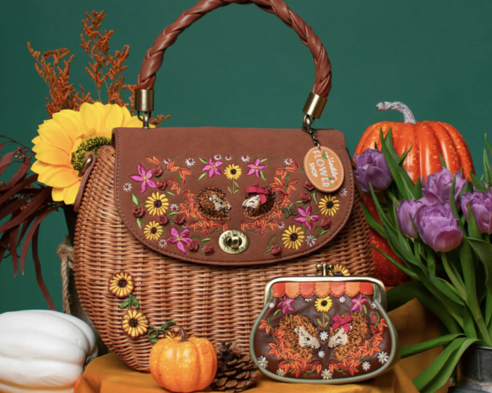 A brown wicker handbag with a twisted top handle and a matching coin purse, both decorated with colorful floral and butterfly embroidery, styled on a yellow fabric surface with autumn props against a dark green background.