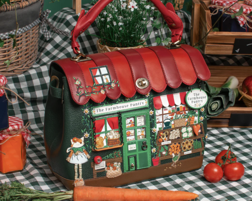 A decorative handbag shaped like a small shop front, with a red handle and a multicoloured illustrated facade, displayed on a green-and-white checkered tablecloth with carrots, tomatoes, baskets, and plants around it.
