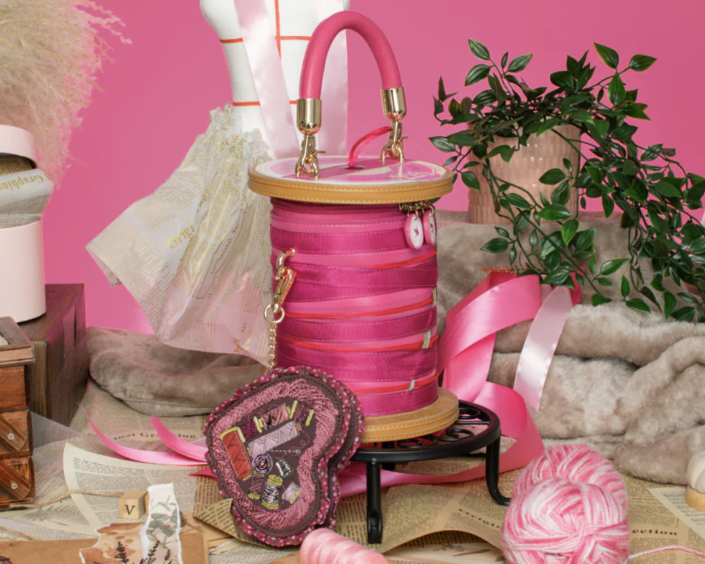 A pink spool-shaped handbag with a top handle, gold-tone hardware, and a quilted floral front, displayed on a small black stand in a styled craft room scene with pink yarn, thread, ribbon, and a mannequin torso against a pink backdrop.