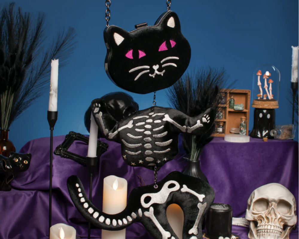 A Halloween-themed tabletop display featuring a black cat-shaped purse with pink eyes, a skeleton cat figurine, candles, a skull, and dark decorative grass against a blue backdrop.