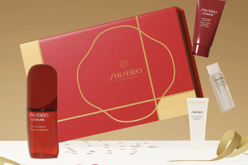 Shiseido gift set featuring a large red box with gold detailing, a red serum bottle, a red hand cream tube, and a small clear bottle, arranged on a white surface with gold ribbons and confetti against a warm beige background.
