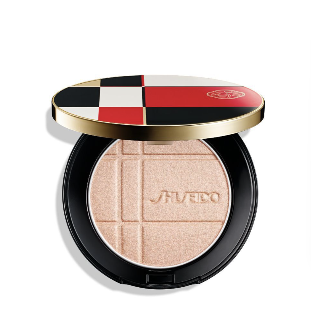 Shiseido color + enahncer Pressed Powder compact shown open, featuring a black circular case with a gold rim and a lid decorated in a red, black, white, and beige checkered pattern. The pressed powder inside is a light, shimmery peach-beige with the Shiseido logo embossed in the center.