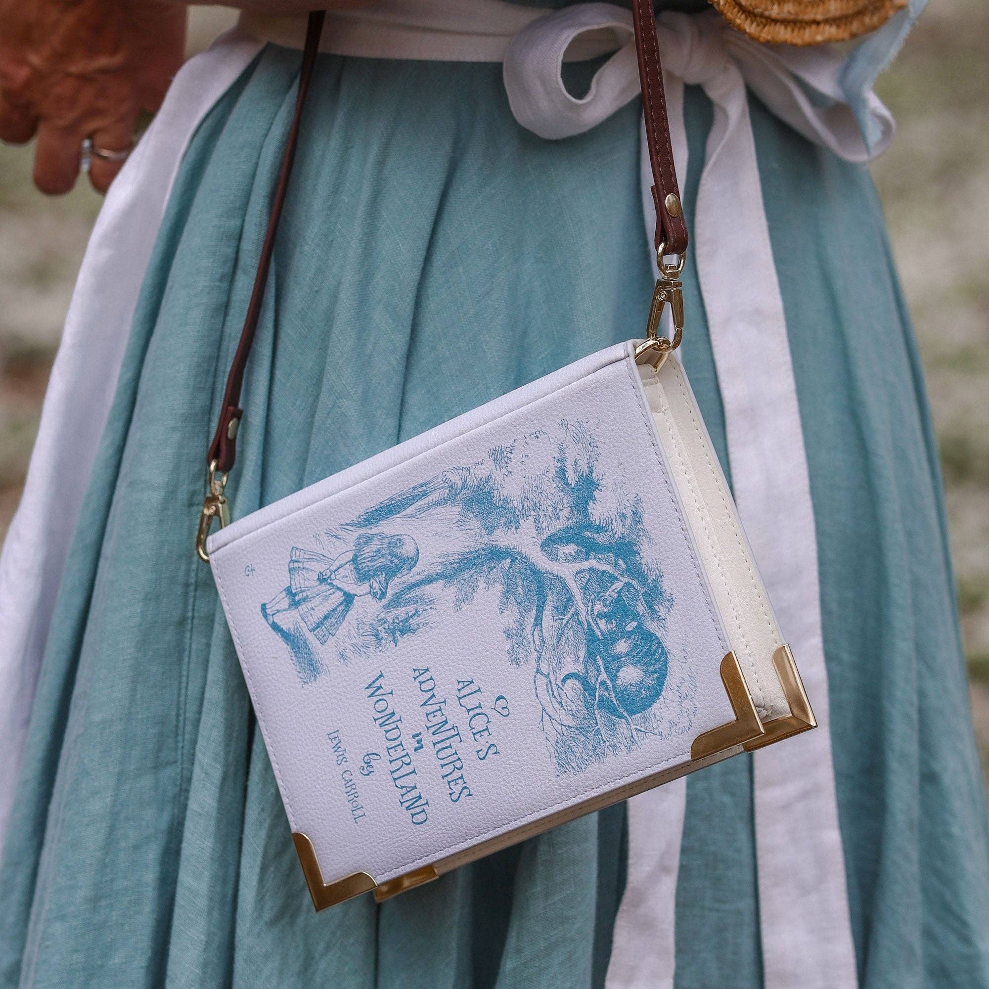 A purple faux-leather crossbody bag designed to look like a vintage Alice in Wonderland book, featuring a dark brown leather shoulder strap and gold-tone hardware.