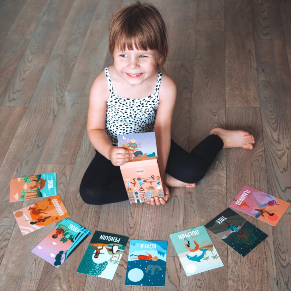 Child sitting on a wooden floor holding a book with various books spread around on the floor.