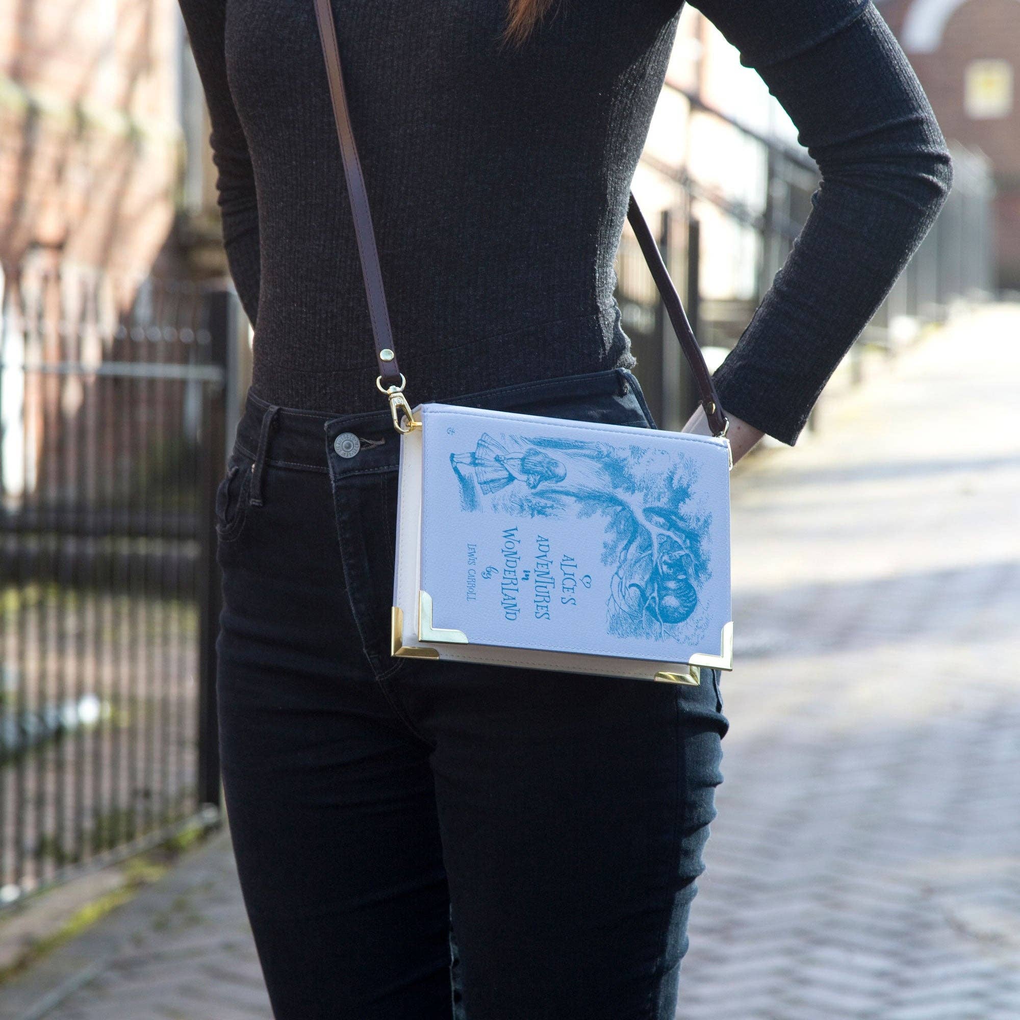A purple faux-leather crossbody bag designed to look like a vintage Alice in Wonderland book, featuring a dark brown leather shoulder strap and gold-tone hardware.