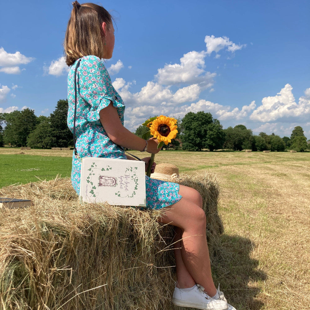 Woman sitting on a hay bale in a field holding a sunflower, with a blue sky and green trees in the background.