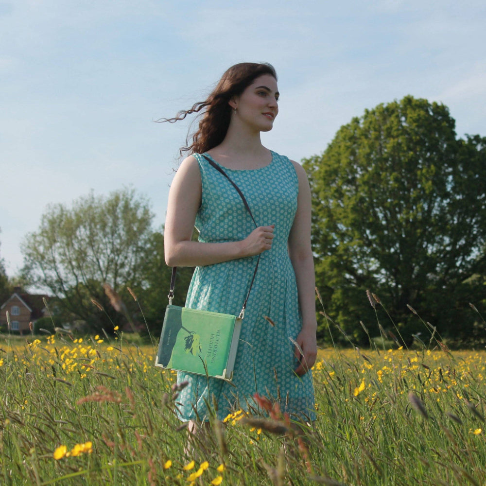 Woman in a green dress holding a green book bag in a field with yellow flowers and trees in the background.