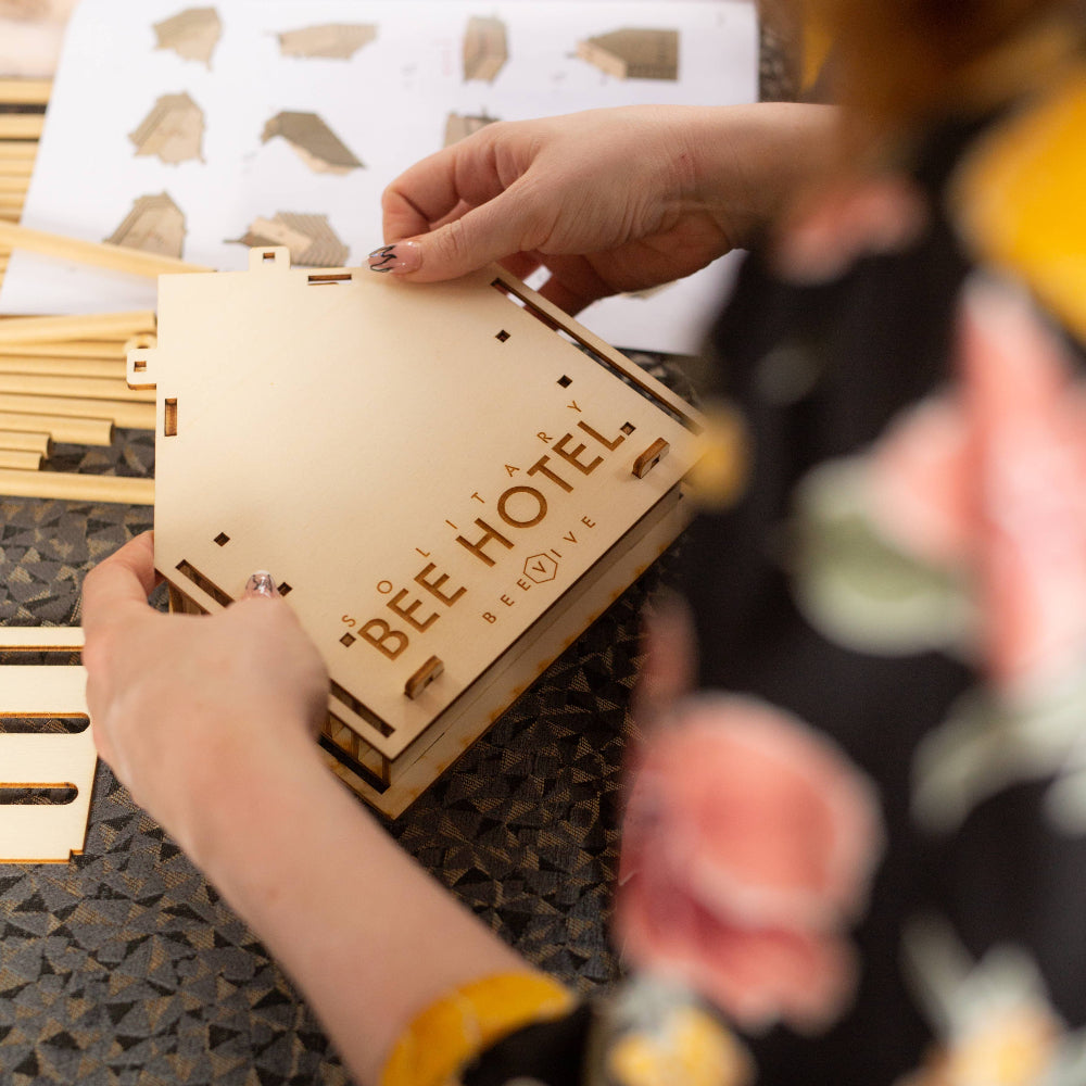 Person assembling a wooden bee hotel with a floral-patterned sleeve in the foreground.