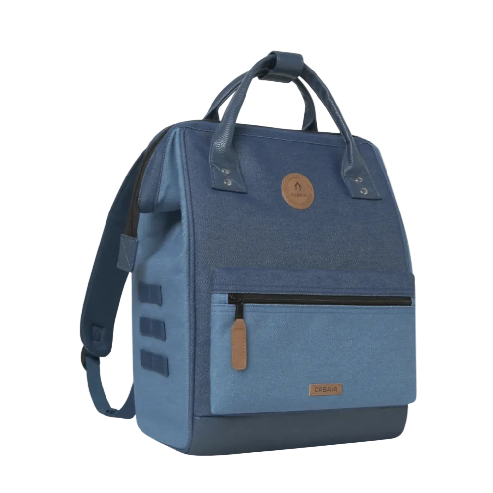 A blue canvas backpack with a dark navy base, featuring a front zippered pocket, top handles, and a circular tan leather logo patch on a light background.