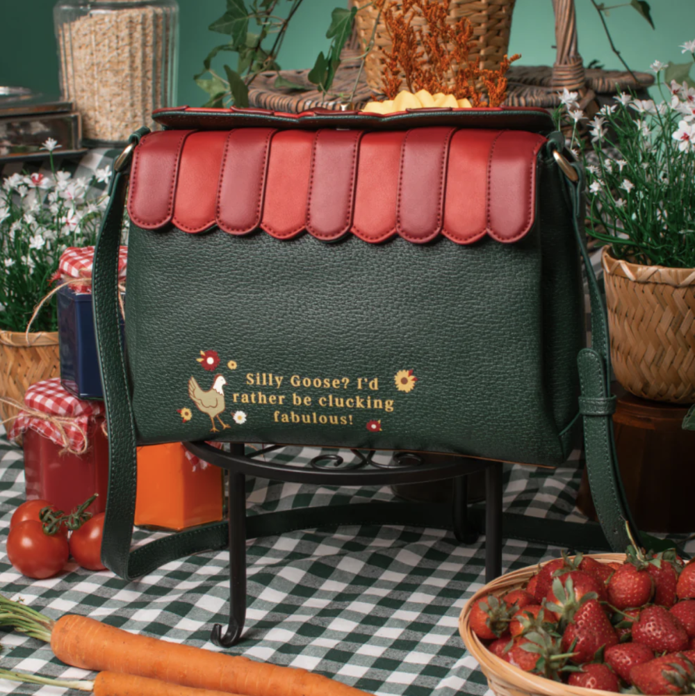 A dark green pebbled leather shoulder bag with a scalloped red top flap, displayed on a black stand in a rustic farm-themed setting with carrots, strawberries, and potted plants.