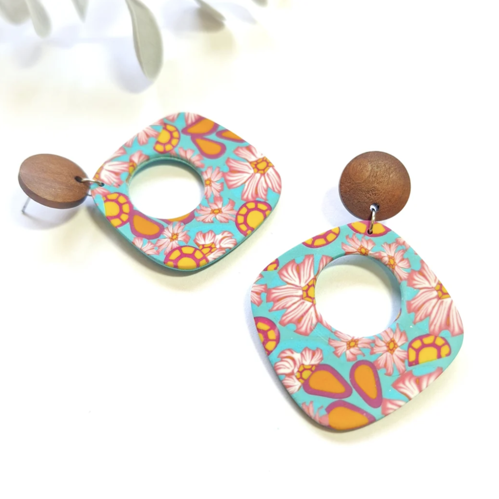 A pair of dangling earrings with irregular square-shaped acrylic pendants featuring a retro floral pattern in turquoise, pink, orange, and yellow, suspended from round wooden studs, displayed on a white background.