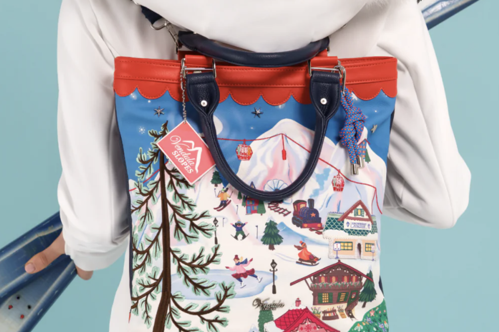 A person wearing a white coat with a fluffy hood, shown from the back while wearing a colorful ski resort backpack. The backpack features a detailed winter mountain scene with cabins, trees, and skiers, accented with red trim and dark blue handles.