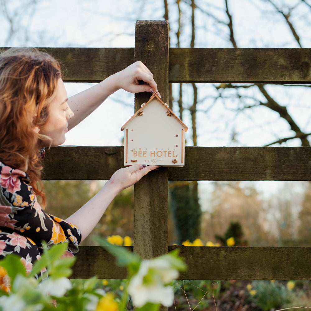 Person hanging a 'Bee Hotel' sign on a wooden fence with flowers and trees in the background