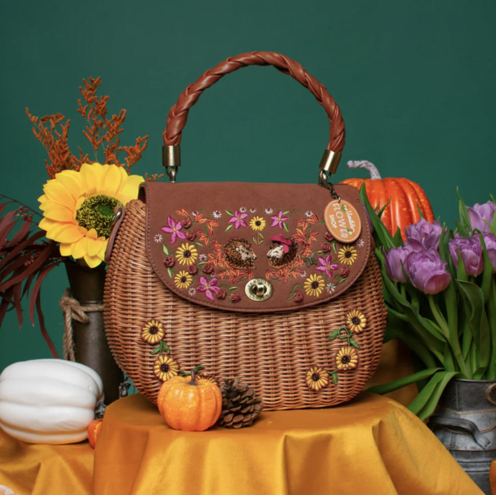 A brown wicker handbag with a braided top handle, decorated with colorful floral embroidery and appliques on the front flap. The bag is styled on a yellow fabric surface with autumn props including pumpkins, tulips, and a sunflower against a dark green background.