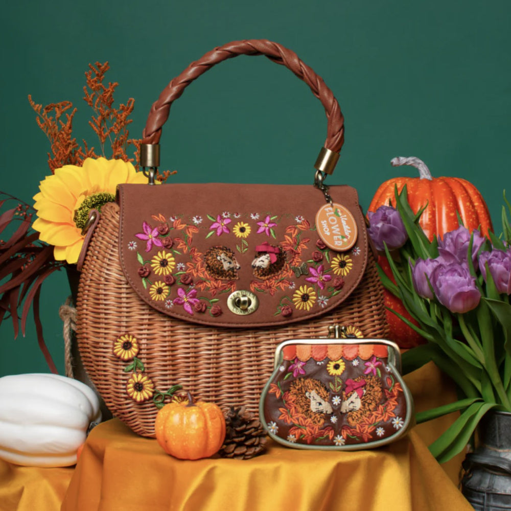 A brown wicker handbag with a twisted top handle and a matching coin purse, both decorated with colorful floral and butterfly embroidery, styled on a yellow fabric surface with autumn props against a dark green background.