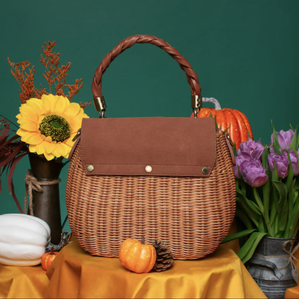 A brown woven wicker handbag with a braided top handle, displayed on a draped yellow fabric surface with autumn props including pumpkins, flowers, and a green backdrop.