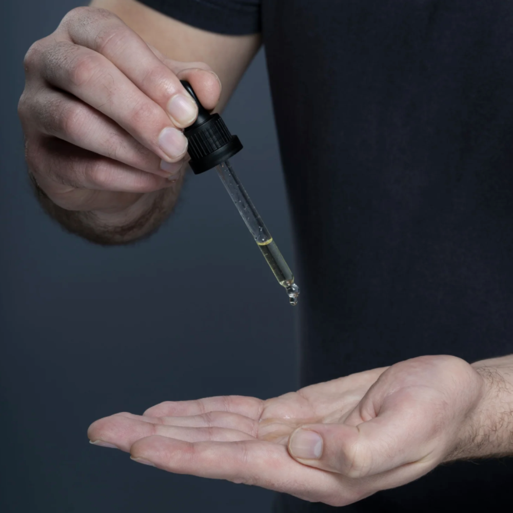 A close-up of a person holding a glass dropper filled with a yellow oil above their open palm, wearing a black t-shirt against a dark gray background.