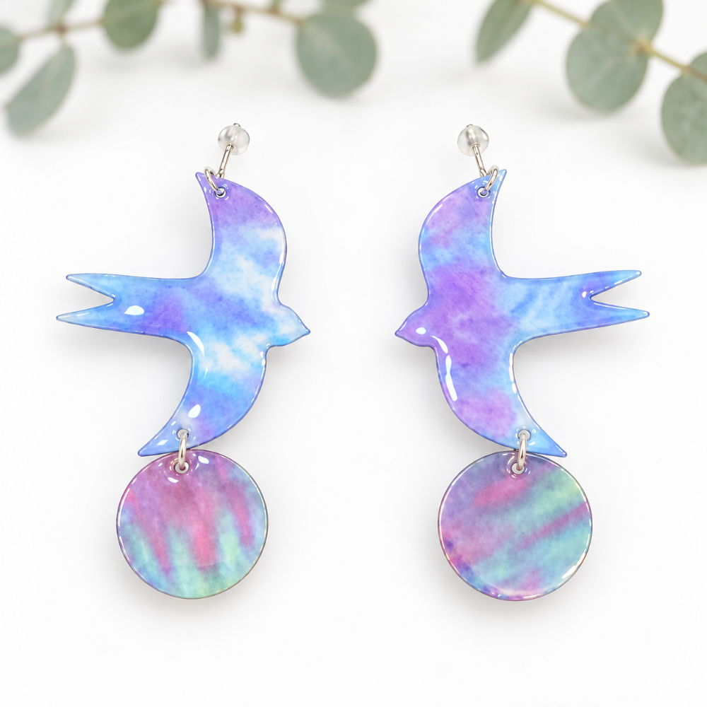 A pair of dangling earrings shaped like birds with circular iridescent shell-like drops, shown on a white background with soft green leaves blurred in the background.