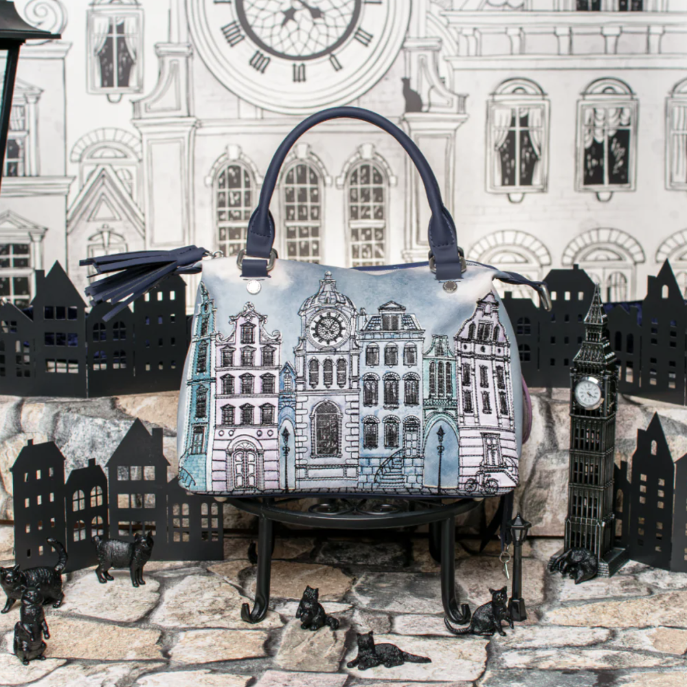 A structured handbag with a printed illustration of London buildings, displayed on a miniature black stand in front of a London-themed backdrop featuring paper cutouts of Big Ben and a clock tower.