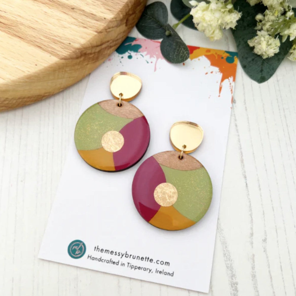Colorful circular earrings on a branded card with a wooden board and flowers in the background.