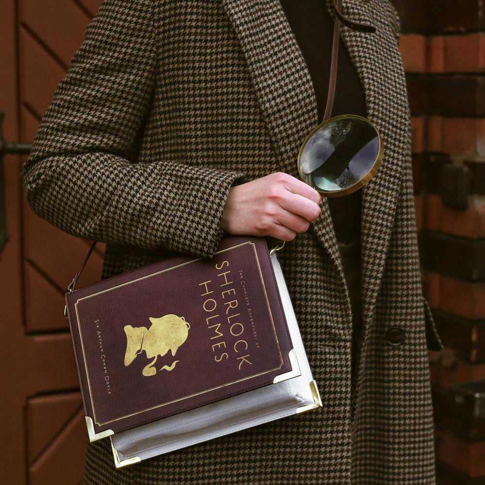 Person in a coat holding a book titled 'Sherlock Holmes' and a magnifying glass against a brick wall.