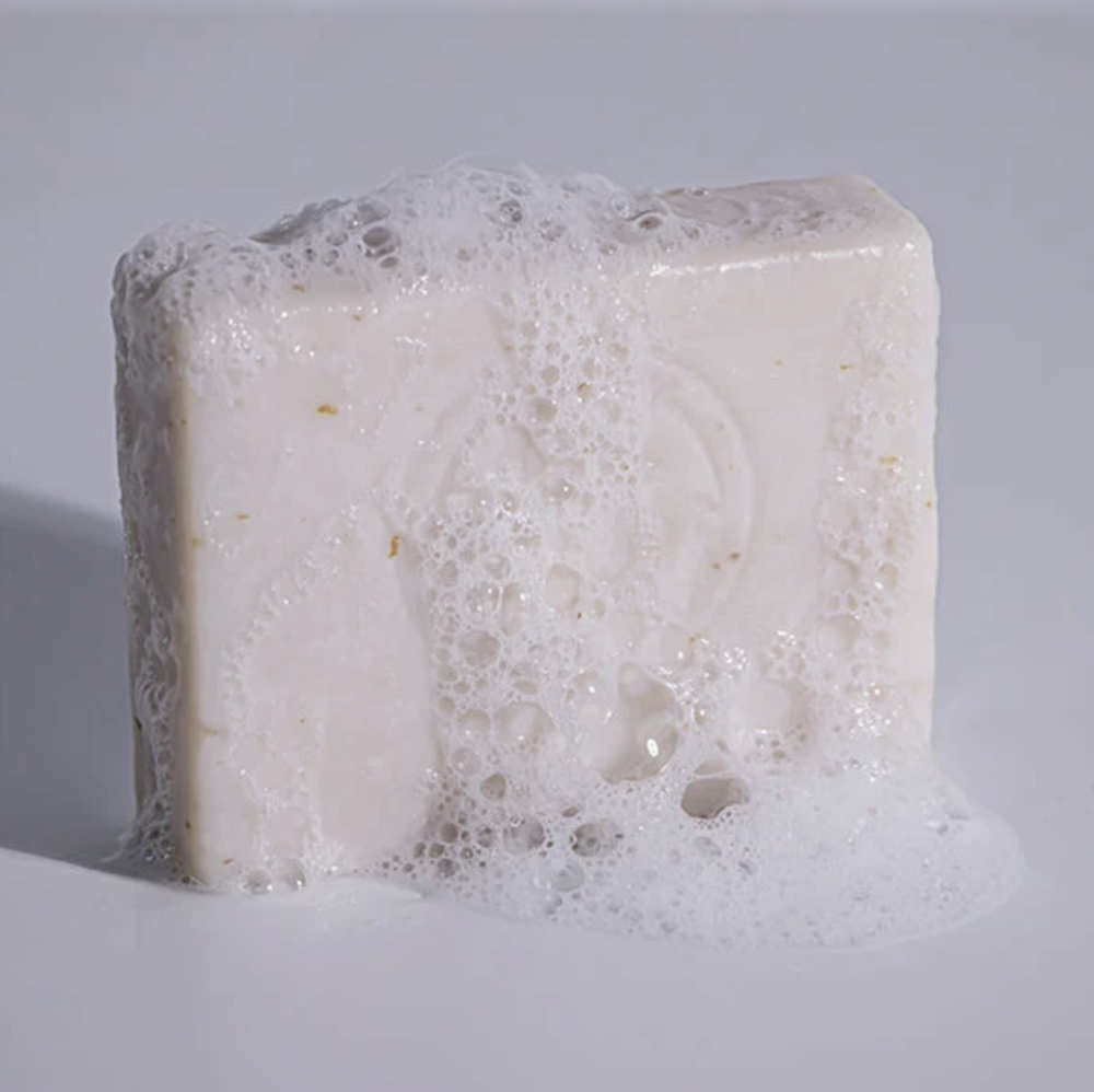 A rectangular bar of creamy off-white soap covered in dense white foam, photographed on a clean light gray background.