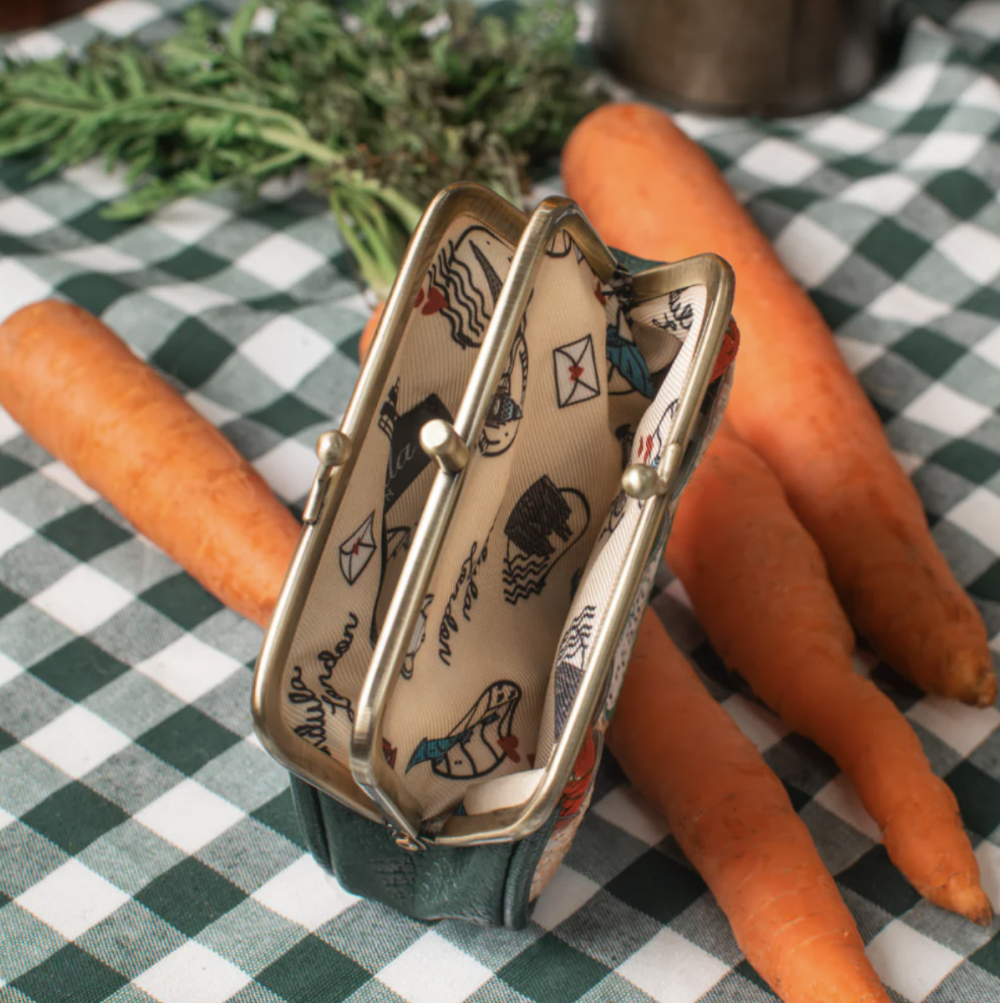 A small kiss-lock coin purse with a cream-colored printed fabric lining and a dark green exterior, styled on a green and white checkered tablecloth with carrots.