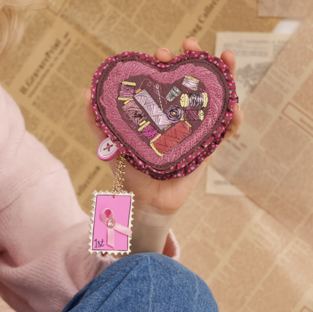 A hand holds a heart-shaped pink and magenta embroidered pouch decorated with sewing-themed patches and a dangling pink tag, set against a background of vintage newspaper pages.