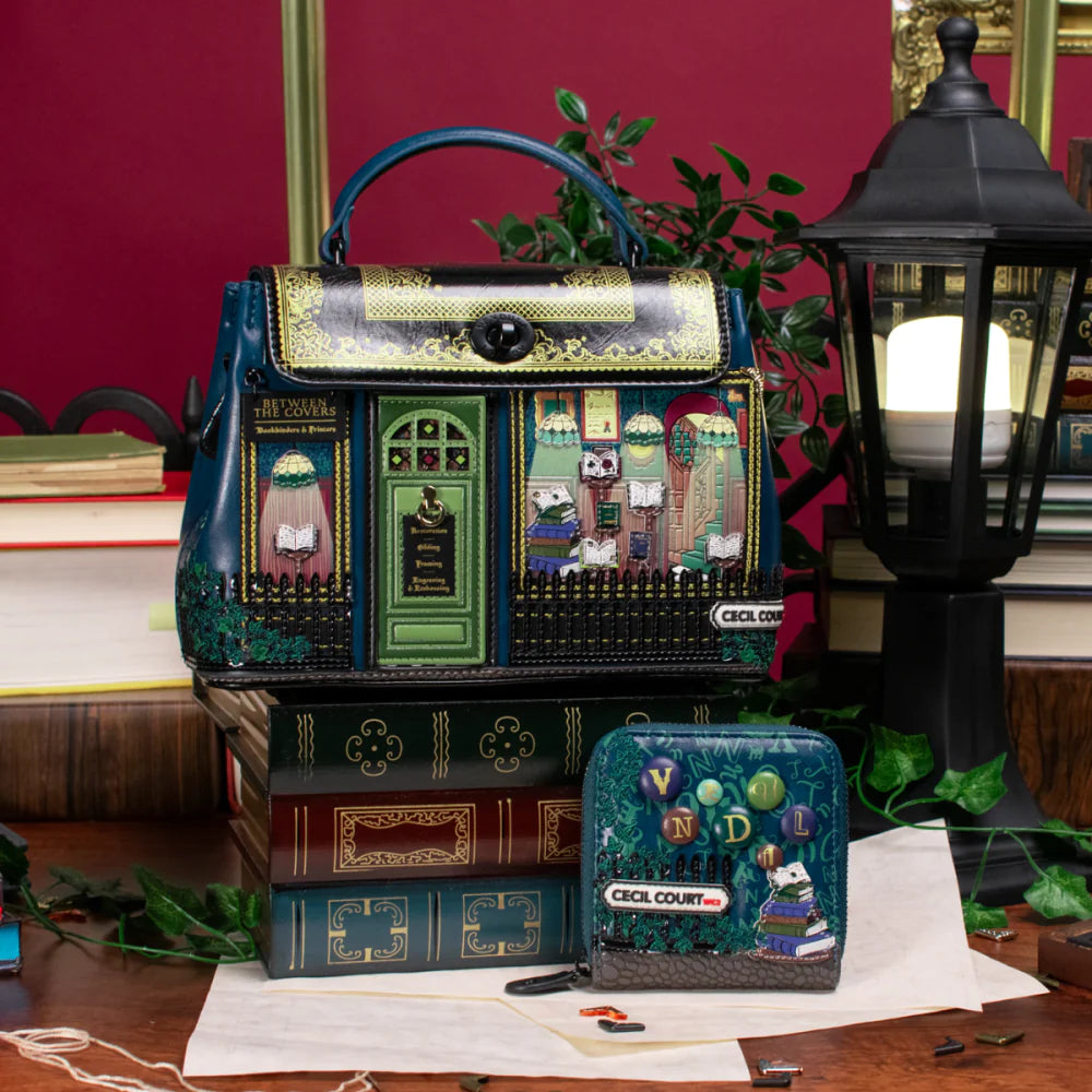 Decorative handbag with book-themed design on a table with books and a lamp.