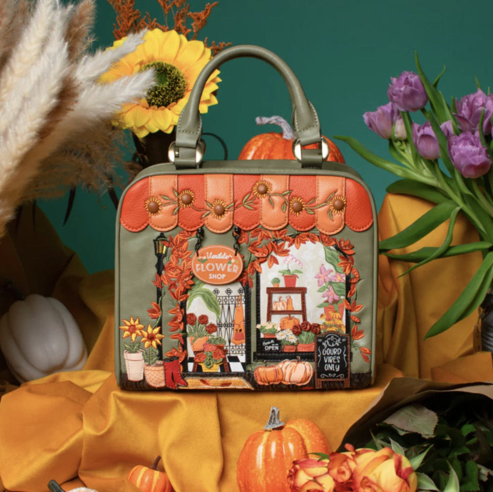 A structured handbag with a warm olive green base and an intricate autumn-themed illustration on the front, styled with pumpkins, flowers, and draped yellow fabric against a teal background.