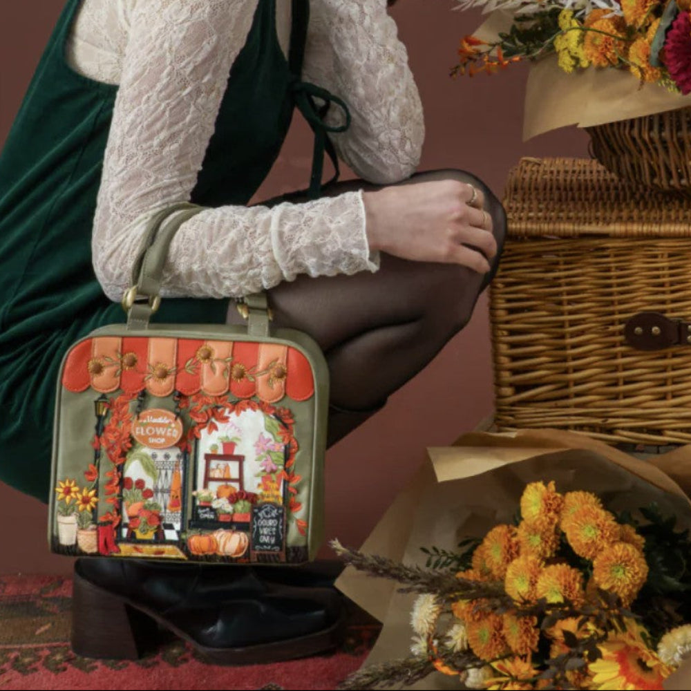 A woman in a dark green dress and white lace top crouches beside a vintage-style illustrated handbag, with wicker baskets and dried flowers arranged around her in a styled indoor scene.