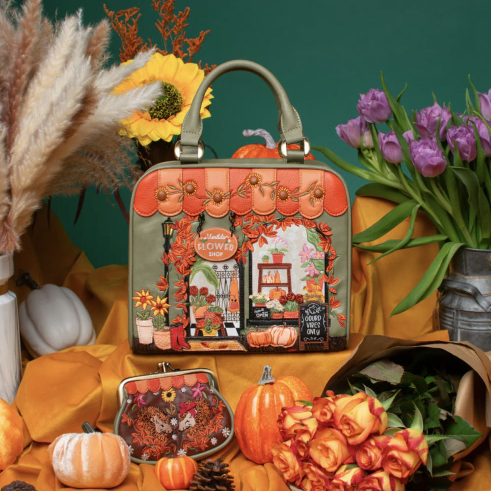 A decorative handbag with an illustrated autumn storefront scene, styled in a fall-themed arrangement with pumpkins, flowers, and draped orange fabric against a dark green background.