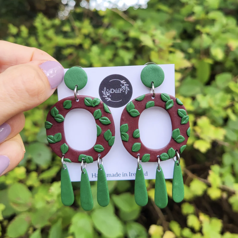 Pair of earrings with green leaves and red circular designs on a card, held against a green leafy background.