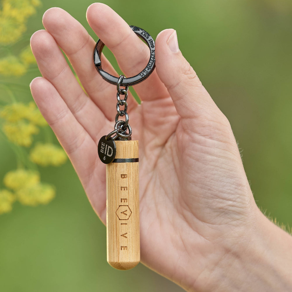 Hand holding a wooden keychain with 'BEEVIVE' branding against a blurred natural background.