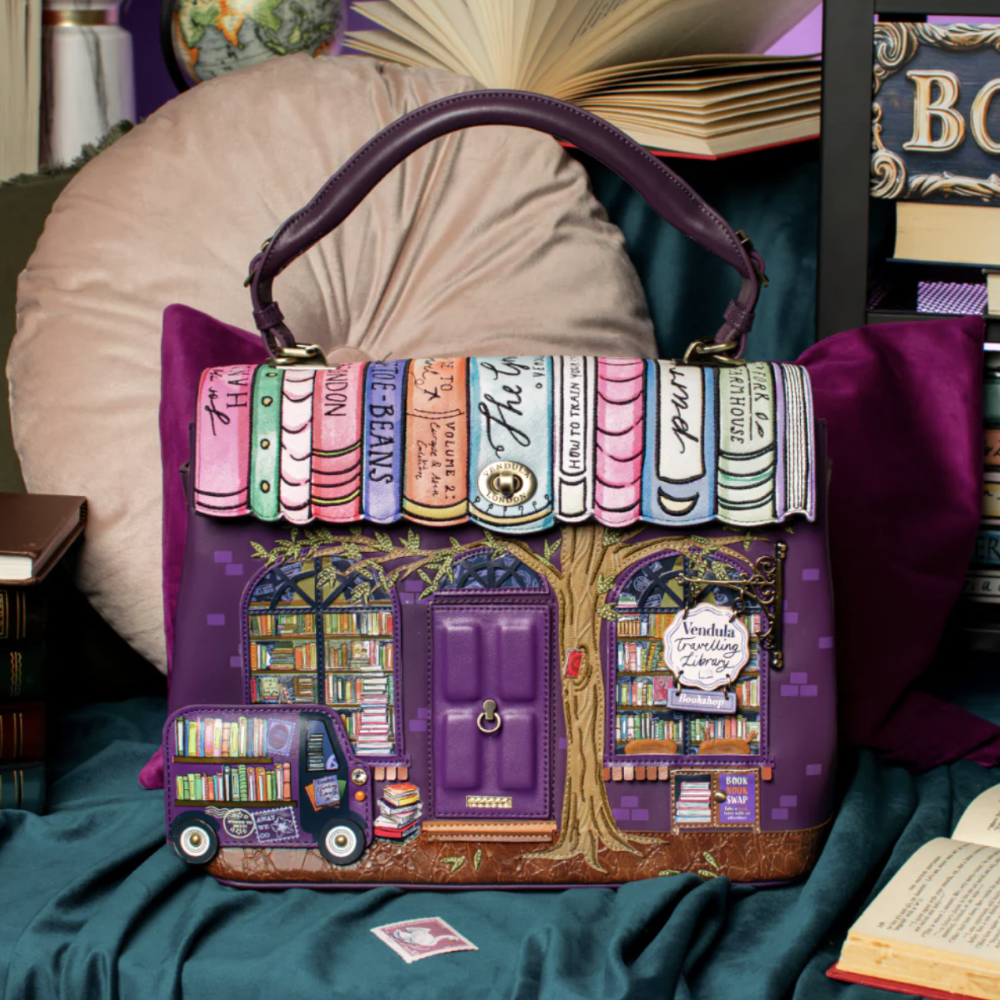 A purple handbag with a whimsical bookstore design, featuring a handle, book spines across the top, illustrated bookshelves, a tree trunk, and a purple truck carrying books, set against a cozy background of books and fabric.