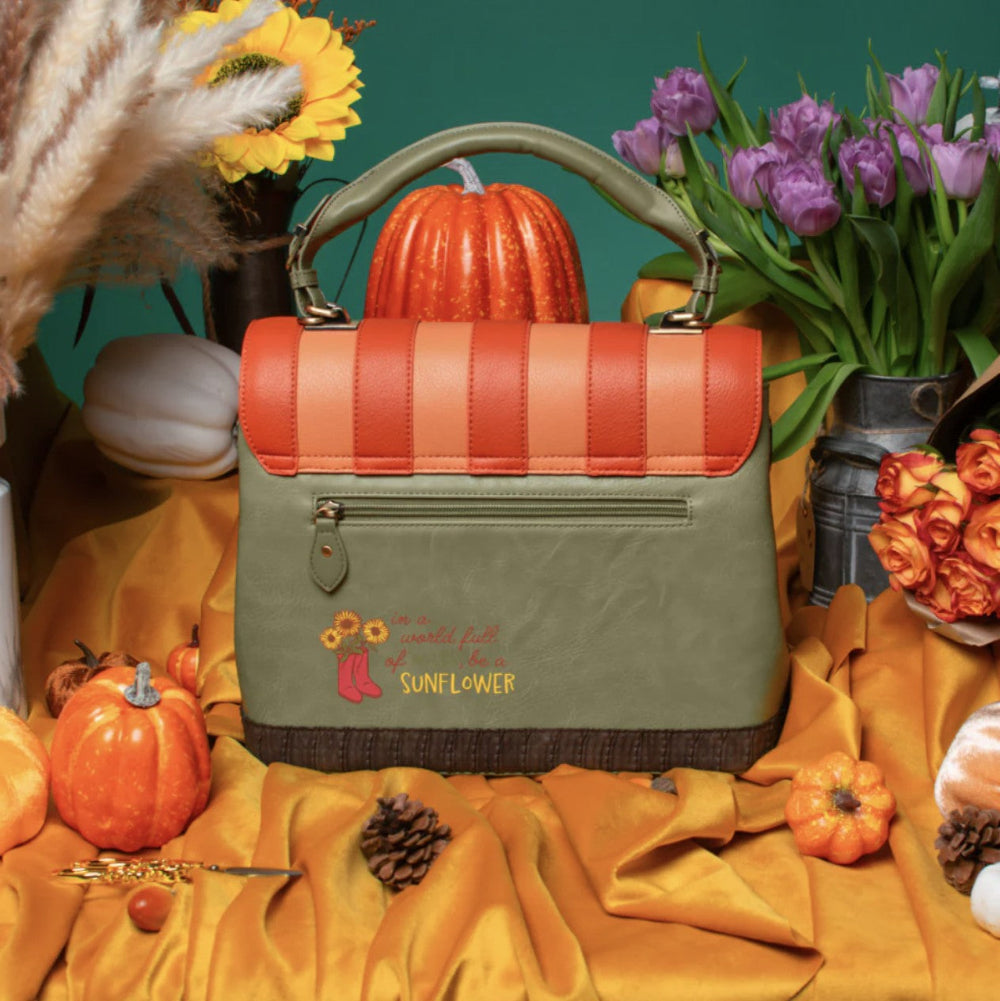 An olive green and orange handbag with a top handle, front zip pocket, and embroidered 'Sunflower' logo, styled on draped orange fabric with pumpkins, flowers, and autumn props against a teal background.