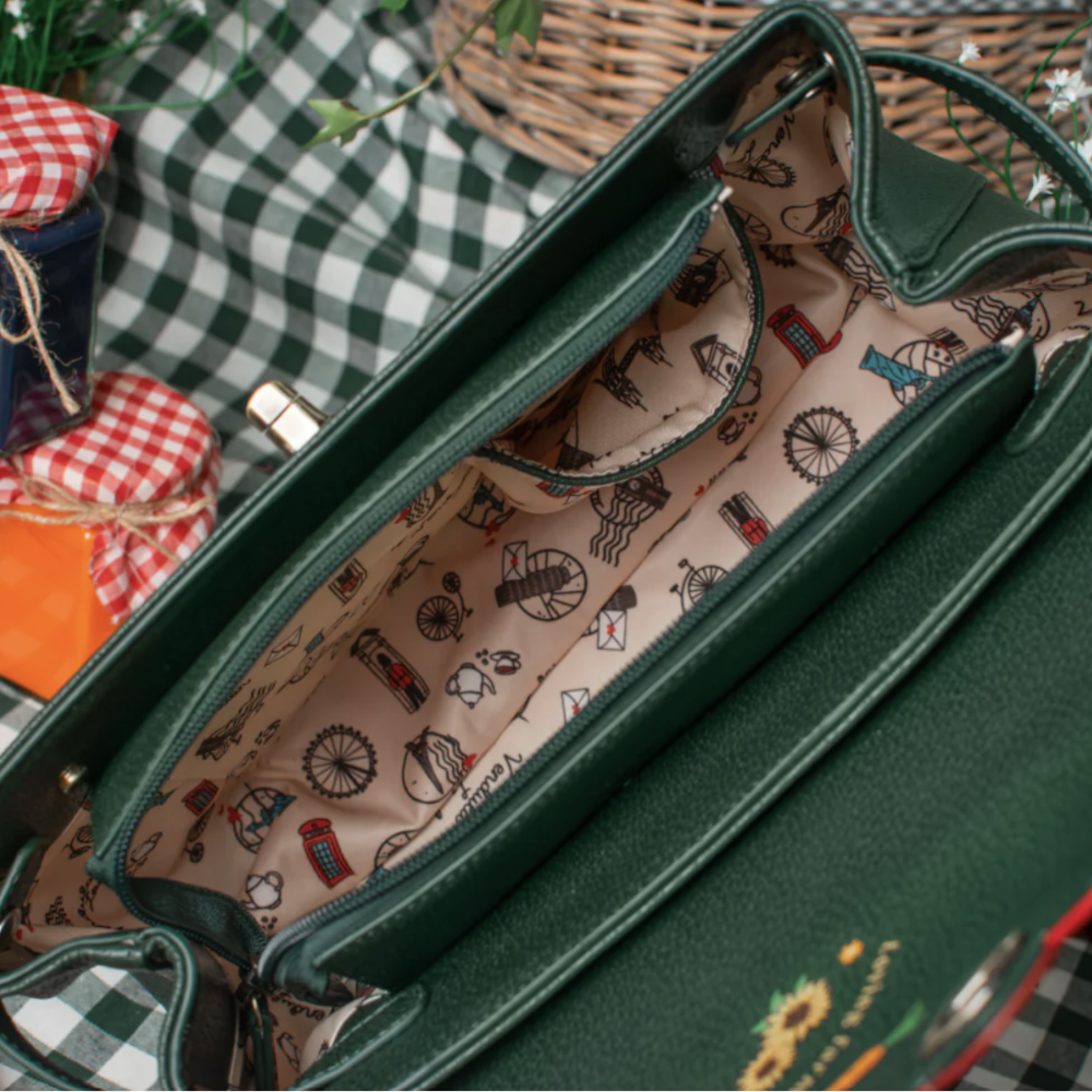 A dark green textured leather handbag shown open, revealing a beige interior printed with vintage red buses, bicycles, and London-themed icons. The bag is styled on a green-and-white checkered picnic blanket with a wicker basket and small fabric bundles in the background.