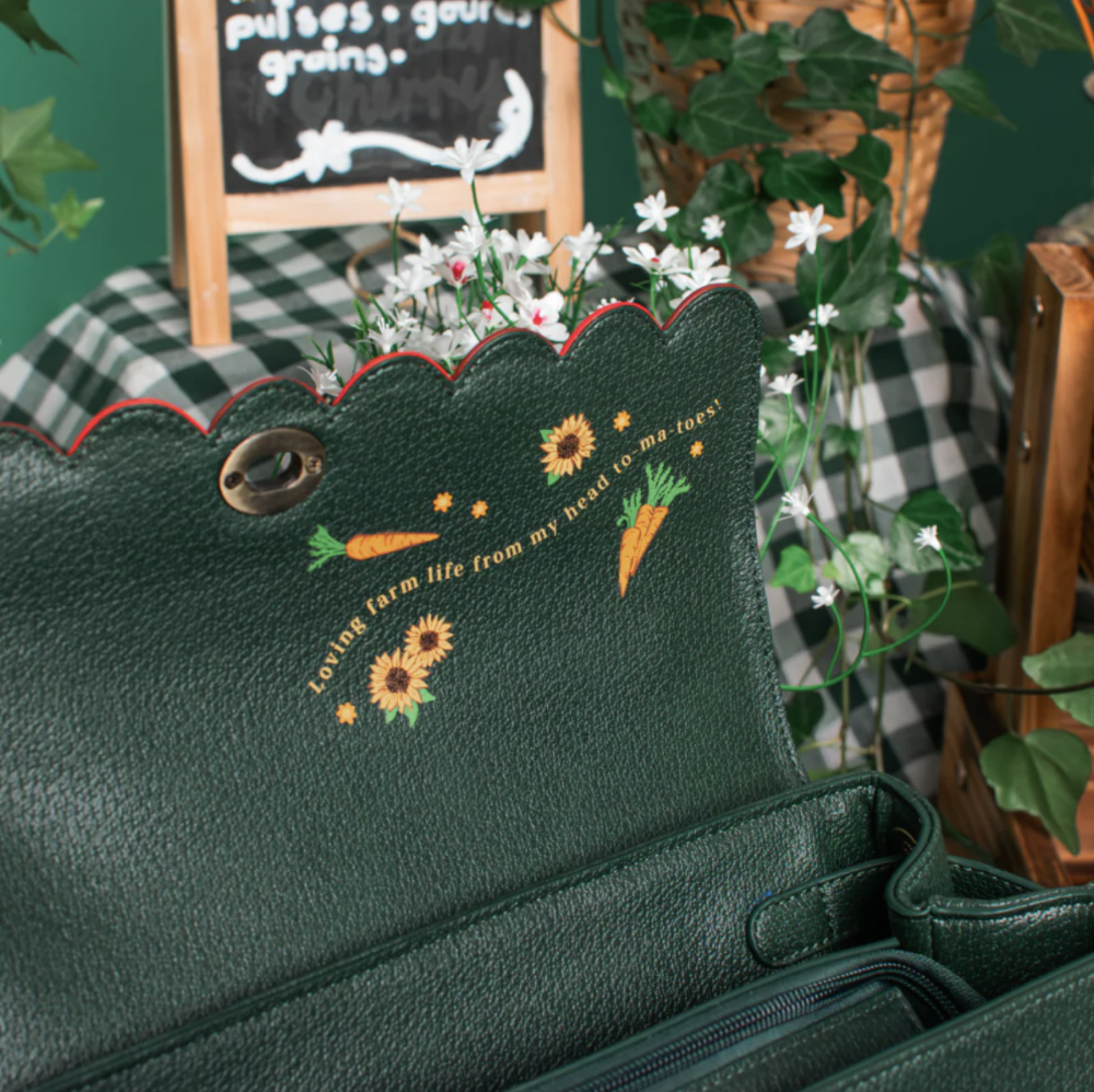 A dark green textured leather clutch bag with scalloped edges, featuring embroidered carrots, sunflowers, and the phrase 'Loving farm life from my head to-ma-toes' on the inside flap. The bag is styled with white flowers on a rustic green and white checkered tablecloth with a chalkboard sign in the background.