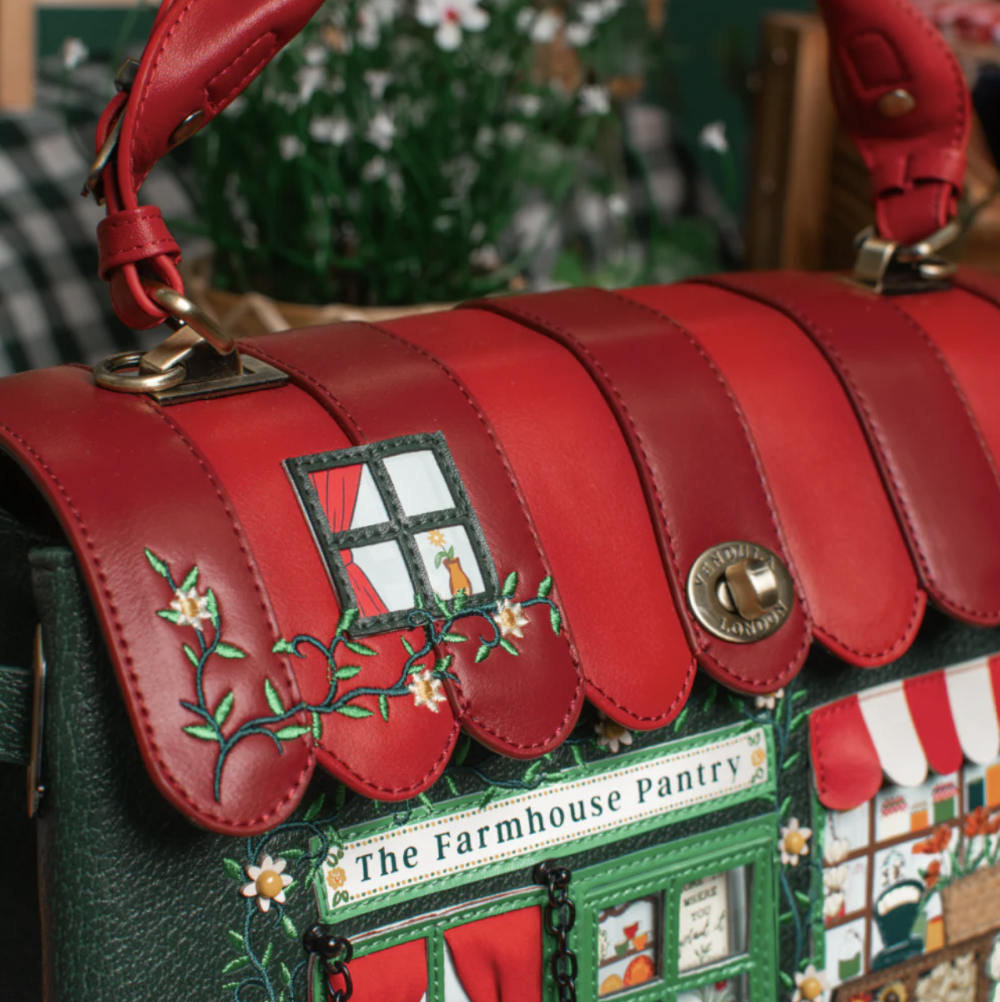 A close-up of a red leather handbag with decorative flap panels, embroidered floral details, and a green side panel featuring a storefront design labeled 'The Farmhouse Pantry.'.