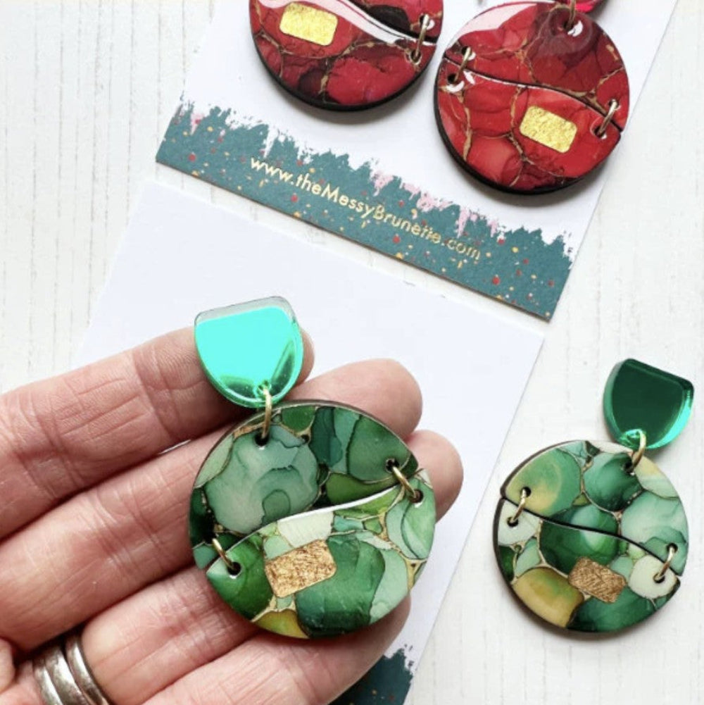 Green and red marbled earrings held by a hand with a brand card in the background.