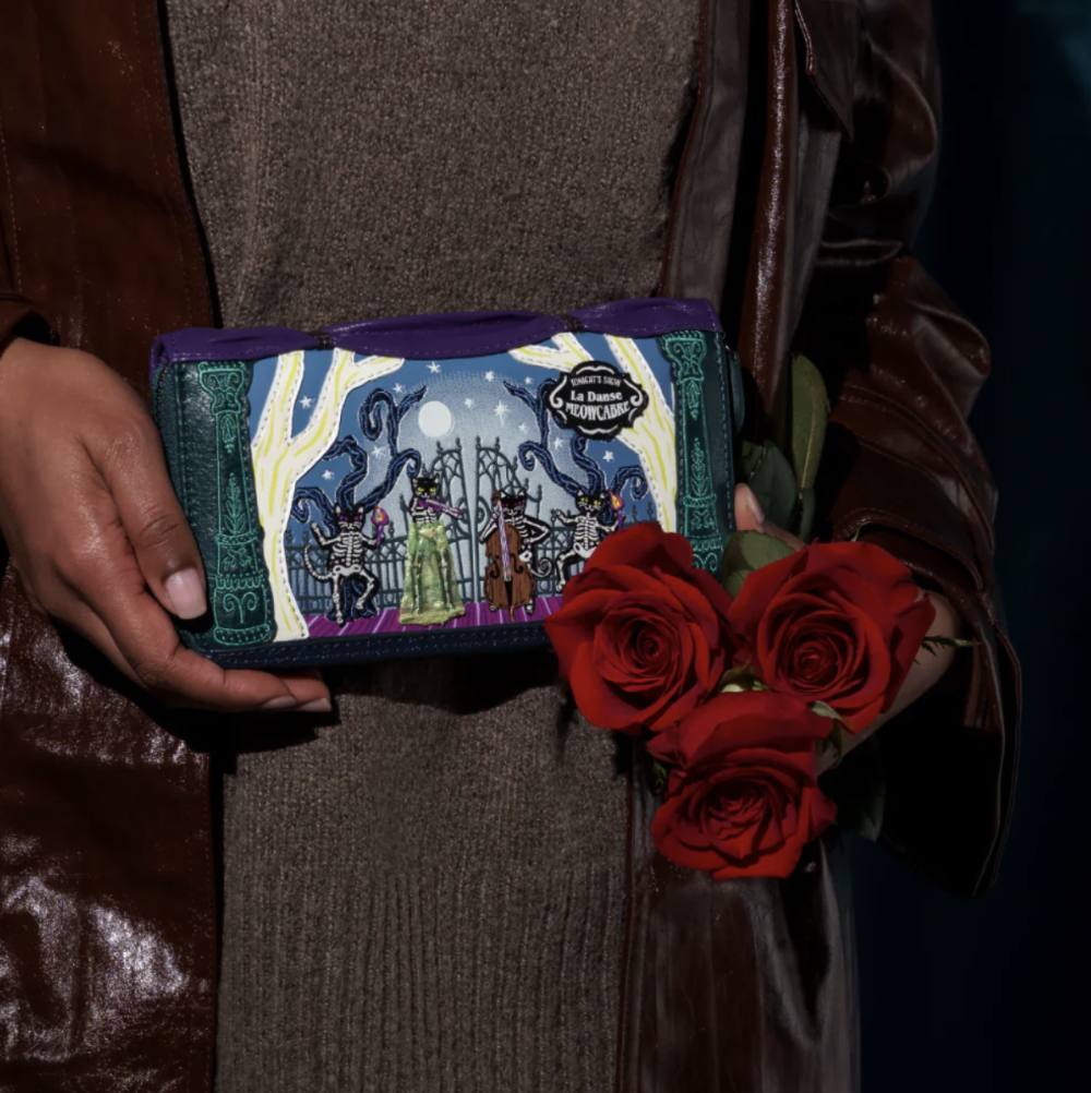 A person in a brown leather jacket and brown knit top holds a small illustrated zip wallet featuring a fantasy scene. The person also holds three red roses against a dark background.