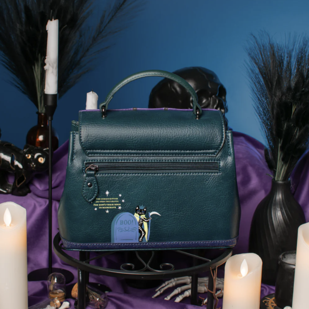A structured teal leather handbag with a top handle, front zip pocket, and a small gothic tombstone charm, displayed on a black stand with candles, purple fabric, and dark decorative props against a blue background.