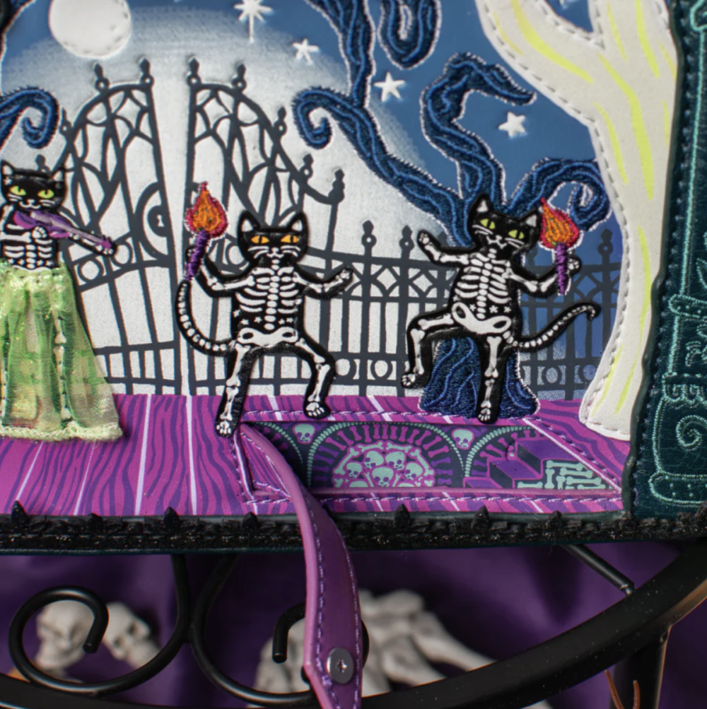 Close-up of a Halloween-themed fabric bag or textile featuring embroidered black cats as skeletons holding torches, a purple gate, a ghost figure in a green dress, and a purple strap hanging in front.