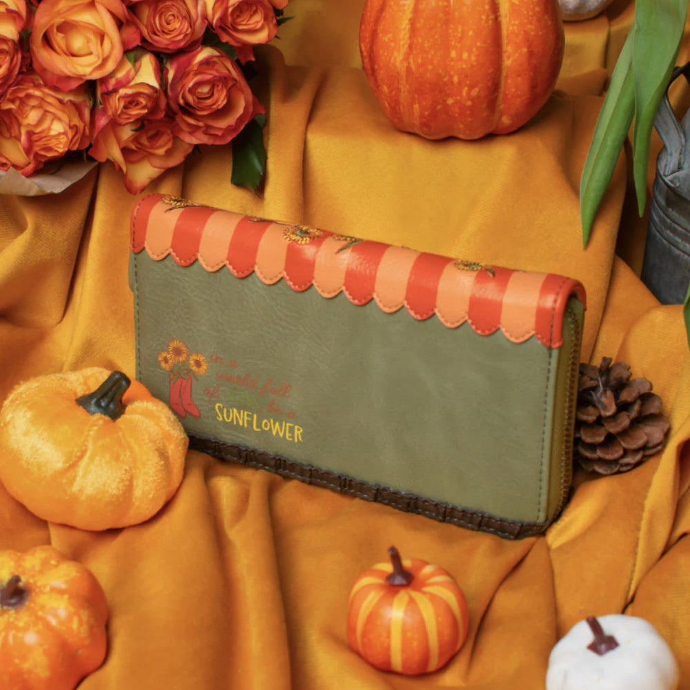 A green wallet with a red and orange striped top border and embroidered text, styled on a draped mustard yellow fabric with pumpkins, roses, and a pinecone.