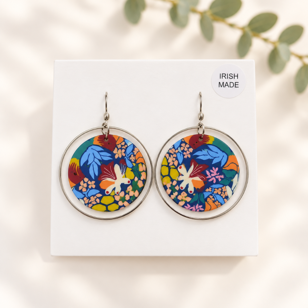 A pair of round silver-tone hoop earrings with colorful floral and bird patterns, displayed on a white card labeled 'IRISH MADE' with soft leaf shadows in the background.