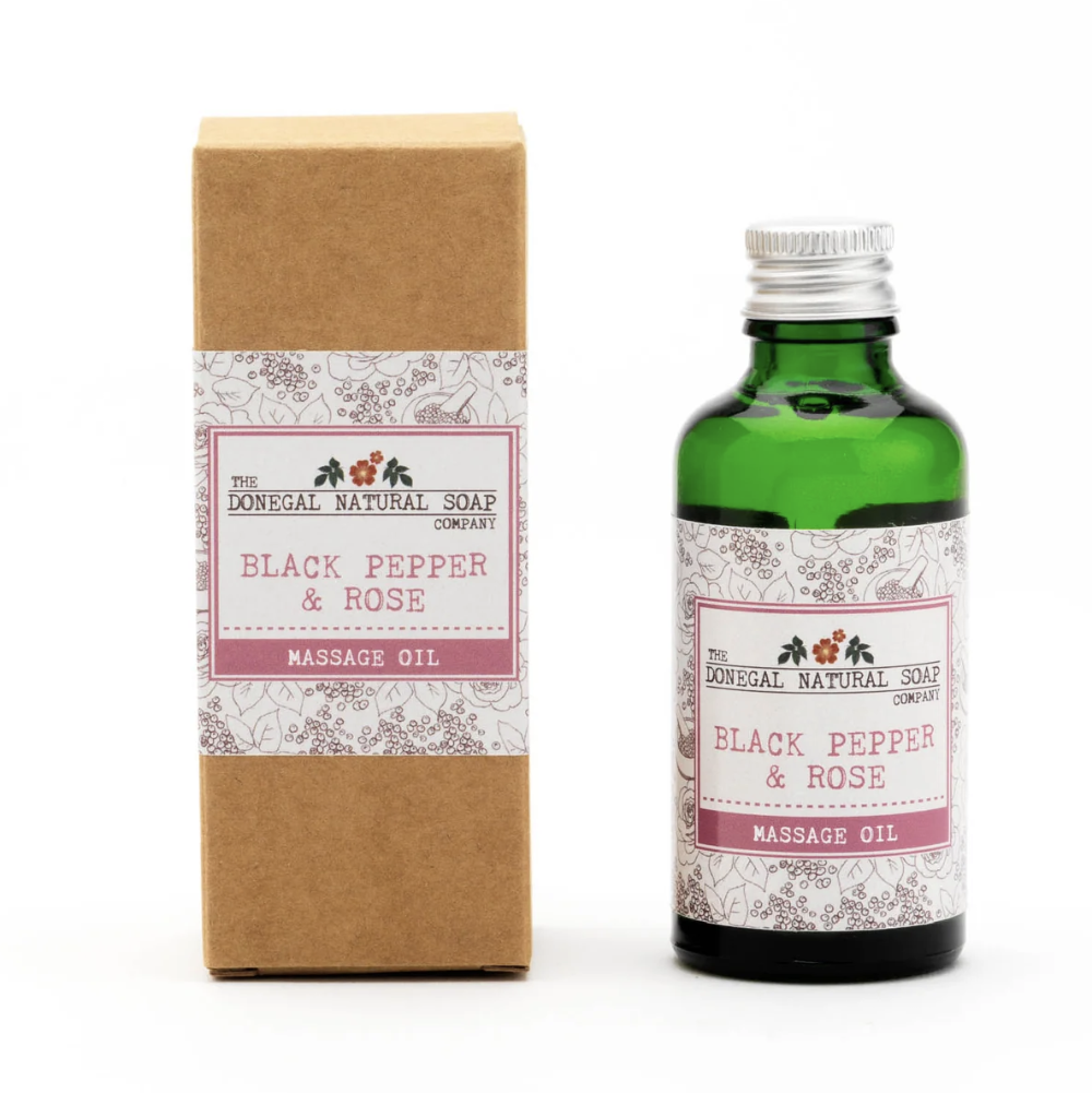 A green glass bottle of massage oil next to its brown kraft paper box packaging, both featuring a white and pink label for 'The Donegal Natural Soap Company' and 'Black Pepper & Rose Massage Oil', on a white background.