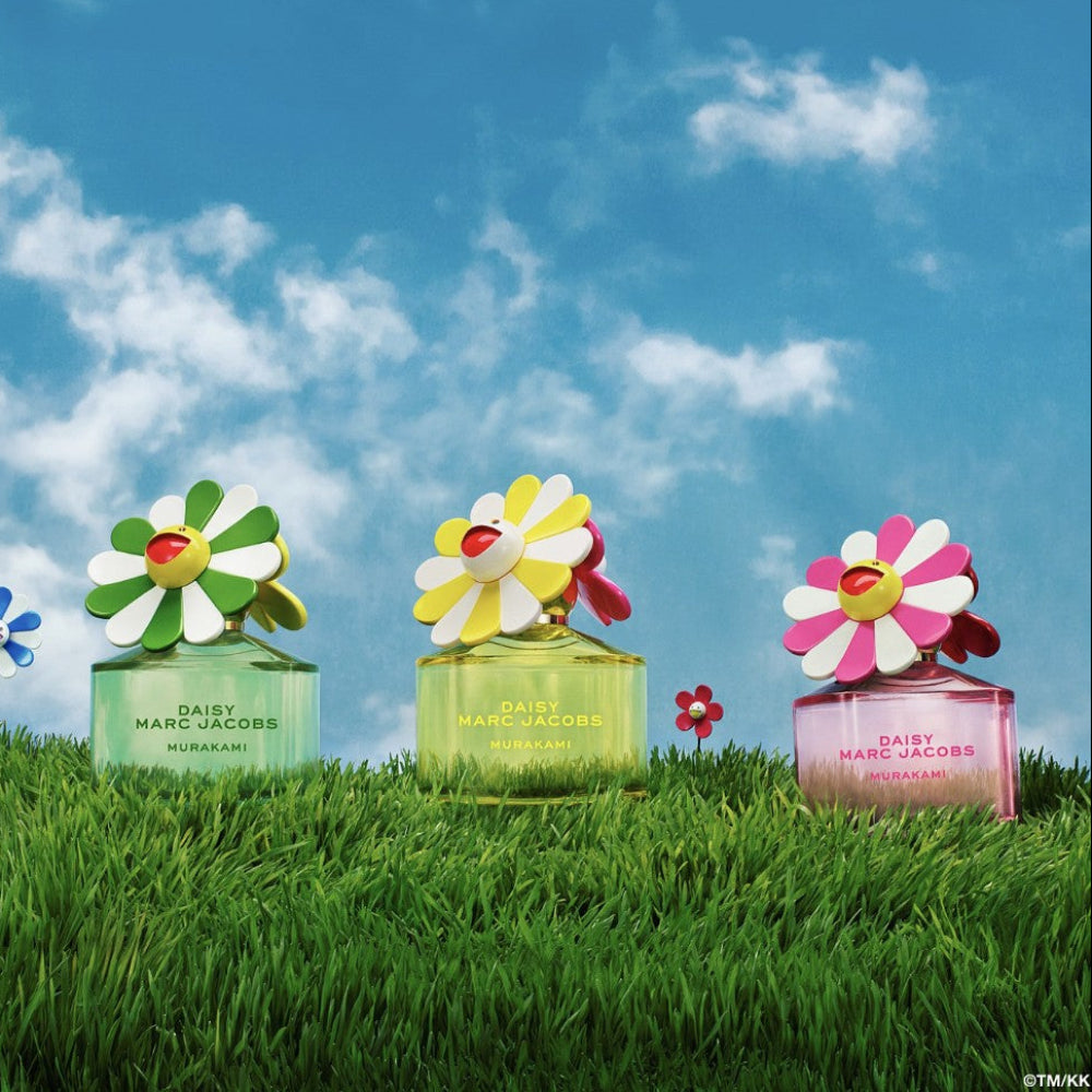 Three Daisy Marc Jacobs perfume bottles with colorful flower designs on a grassy field with a blue sky.