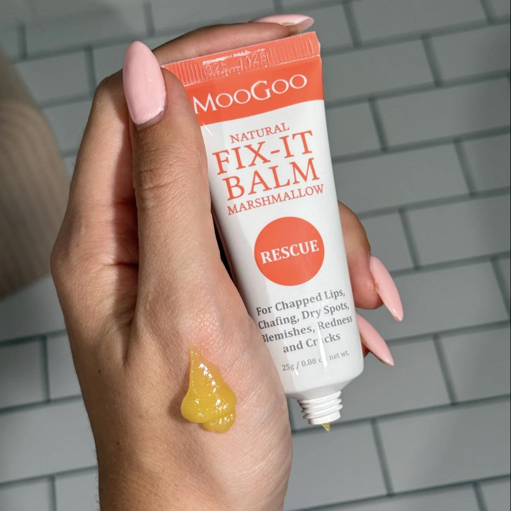 A hand holds a tube of MooGoo Natural Fix-It Balm, with a dollop of yellow balm on the back of the hand against a gray tile background.
