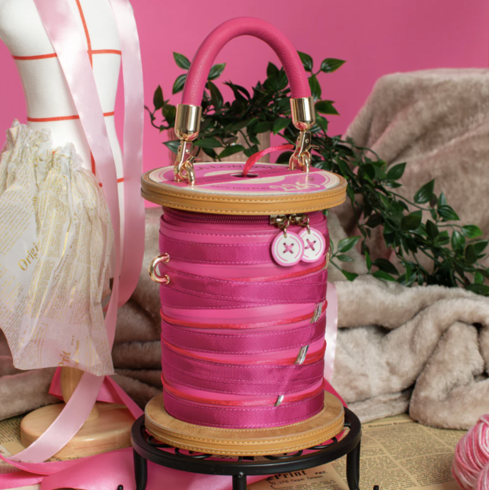 A pink spool-shaped handbag with a top handle, gold-tone hardware, and decorative buttons, styled on a small black stand against a pink background with mannequins, ribbon, and fabric props.