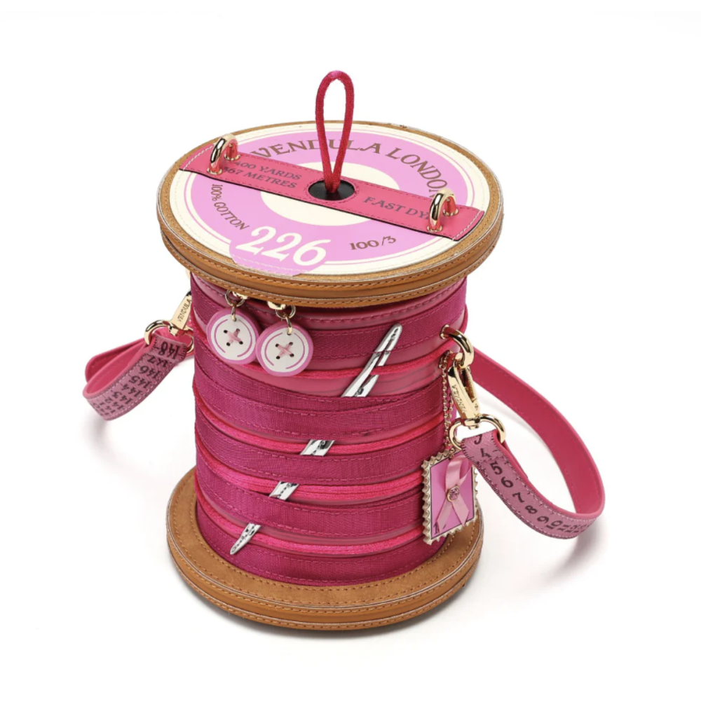 A pink spool-shaped handbag with faux leather stitching, silver zipper details, decorative buttons, and a hanging charm, photographed on a white background.